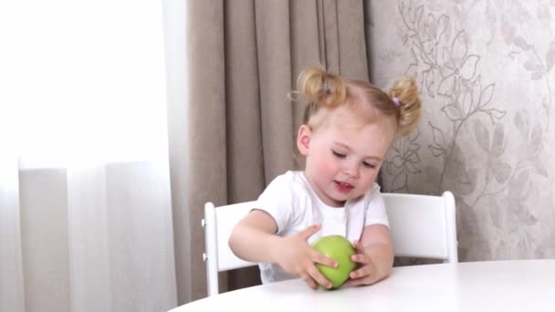 白いTシャツの女の子がテーブルに座って緑のリンゴと遊ぶ — ストック動画
