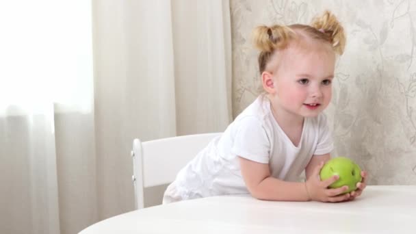 一个穿着白色T恤的小女孩坐在桌旁玩着一个绿色的苹果 — 图库视频影像