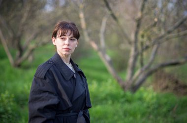 İlkbaharda, siyah yağmurluklu bir kız ağaçların ve yeşil çimenlerin arkasında poz verir..