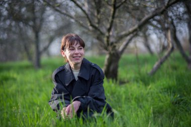 İlkbaharda, siyah yağmurluklu bir kız ağaçların ve yeşil çimenlerin arkasında poz verir..