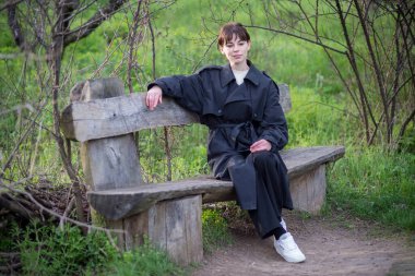 İlkbaharda, siyah yağmurluklu bir kız ağaçların ve yeşil çimenlerin arkasında poz verir..