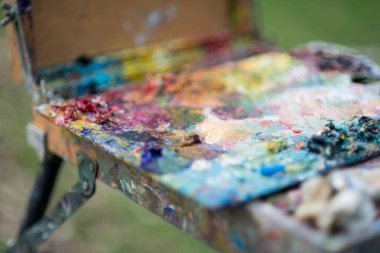 Bir sanatçı baharda parkta bir resim çizer, elleri ve kıyafetleri boyalıdır. Boyaları karıştırmak için bir palet.