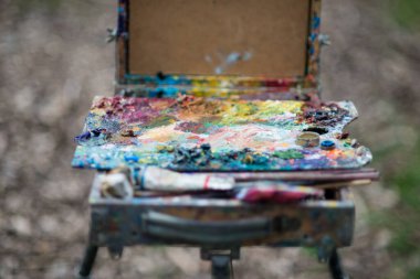 Bir sanatçı baharda parkta bir resim çizer, elleri ve kıyafetleri boyalıdır. Boyaları karıştırmak için bir palet.