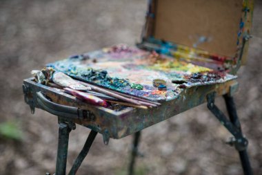 Bir sanatçı baharda parkta bir resim çizer, elleri ve kıyafetleri boyalıdır. Boyaları karıştırmak için bir palet.