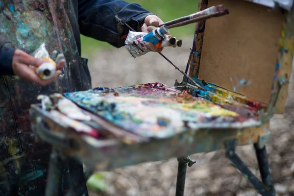 An artist paints a picture in the park in spring, her hands and clothes in paint. A palette for mixing paints.