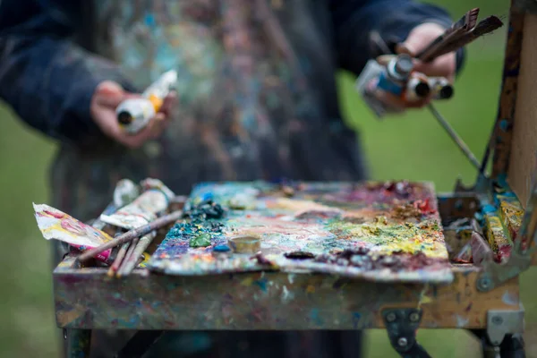 An artist paints a picture in the park in spring, her hands and clothes in paint. A palette for mixing paints.