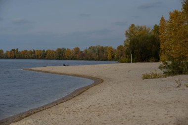 Güz, altın ve kırmızı renkli ağaçlarıyla, nehir kıyısı boyunca sahneyi çerçeveler, ahşap bir çit manzaraya kırsal bir cazibe ekler, gözleri yeşilliklerin arasına yerleştirilmiş tuhaf bir kulübeye götürür..