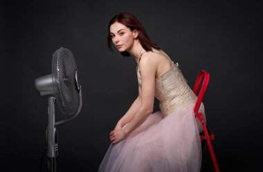 Çekici bir kız, bir fanla fotoğraf stüdyosunda poz veriyor, saçları rüzgarda uçuşuyor, dinamik ve büyüleyici bir sahne yaratıyor, güzelliğini ve zarafetini ön plana çıkarıyor..