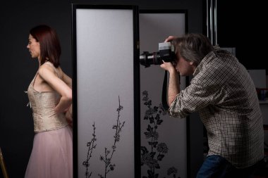 Çekici bir kız aynanın yanındaki fotoğraf stüdyosunda poz verirken, bir fotoğrafçı onun resmini çekiyor, güzellik, zarafet ve profesyonel sanatla dolu bir sahne yaratıyor..