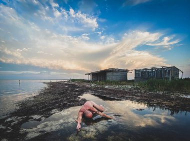 Suda çıplak figür, bulutlu gökyüzü, kırsal kıyı, siyah ve beyaz.