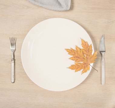 On the table is a white plate with a maple leaf, a fork, a knife and a napkin. A service for lunch for one person