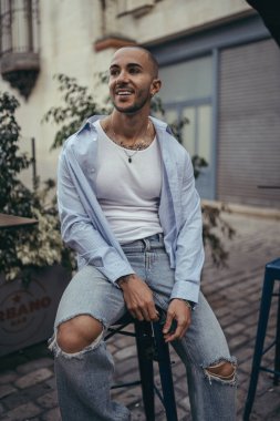 Sandalyede oturan yakışıklı bir gencin portresi. Koyu renk saçlı, beyaz gömlek ve kot pantolon giyen genç adam..