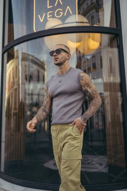 Güneş gözlüklü ve kot pantolonlu yakışıklı genç bir adam şehir merkezinde poz veriyor.