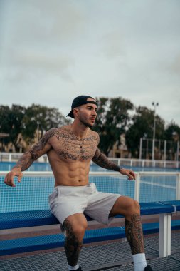 Parkta oturan, spor şapkalı dövmeli yakışıklı adam.