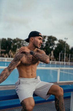 Dövmeli genç kaslı adam spor salonunda oturuyor..