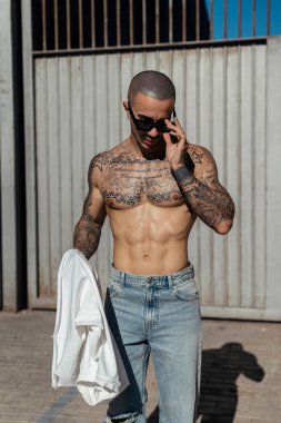 Dövmeli, beyaz tişörtlü, sakallı, başında siyah gömlekli bir adam..