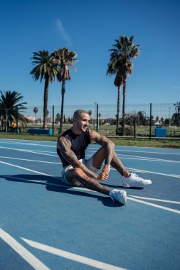 Beyaz tişörtlü ve siyah şortlu kaslı genç bir adam raylarda oturuyor ve mesafeye bakıyor.