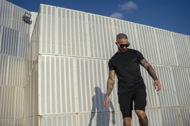 Siyah güneş gözlüklü ve tişörtlü genç kaslı adam bir yaz günü şehirde duruyor..