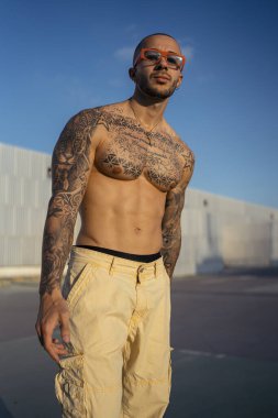 Dövmeli ve güneş gözlüklü genç kaslı adam şehrin arka planında poz veriyor..
