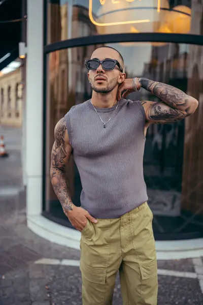 Güneş gözlüklü ve kotlu şık bir gencin binanın yanında poz verdiği portresi.