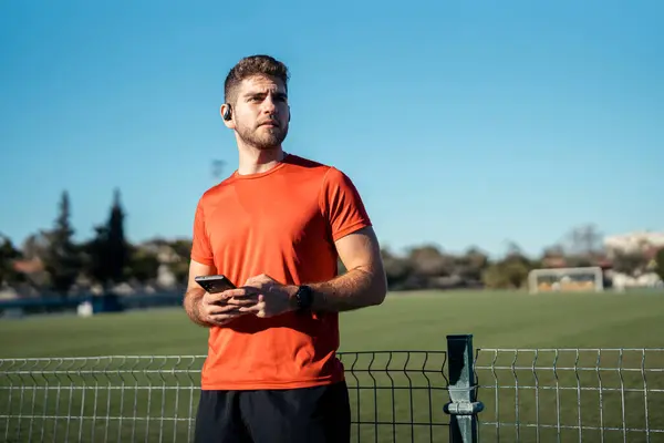 Spor kıyafetleri içinde yakışıklı genç bir erkek sporcu ve kulaklıklarla güneşli bir günde şehir parkında dikiliyor..