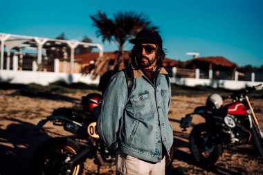 Yol kenarında motosikletli, güneş gözlüklü ve şapkalı sakallı motorcu.