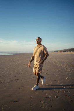 Spor giyimli genç Afro-Amerikan adam kumsalda yürüyor.