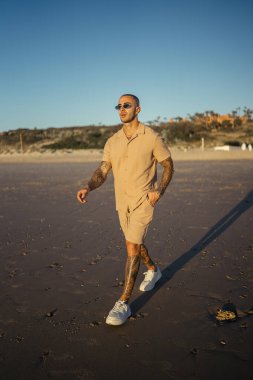 Güneş gözlüklü ve şortlu genç adam sahilde yürüyor.