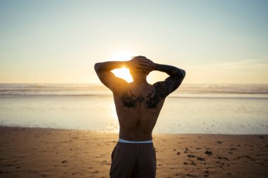 Sahilde kollarını açmış dövmeli bir adamın dikiz görüntüsü.