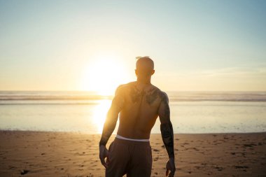 Genç adam gün doğumunda kumlu sahilde duruyor.