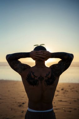 Siyah kapüşonlu ve şapkalı adam sahilde duruyor.