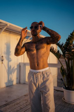 Güneş gözlüklü, şortlu ve elinde güneş doğarken sahilde dövmesi olan yakışıklı Afro-Amerikan bir adam. yüksek kaliteli fotoğraf