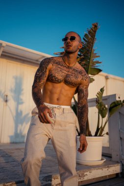 Parkta poz vermiş, kot pantolon ve güneş gözlüğü takan yakışıklı genç bir adam.