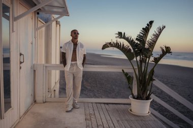 Beyaz pantolonlu, güneş gözlüklü genç bir adam otelin balkonunda şık bir beyaz gömlek giyiyor..