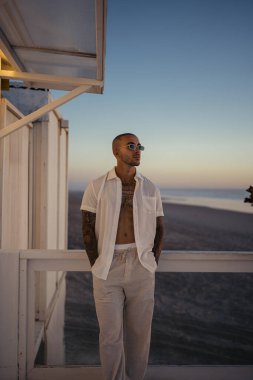 Güneş gözlüklü, kel ve şık giyimli genç bir adamın portresi. Deniz batımının arka planında duran beyaz gömlekli..