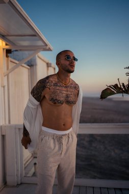 Lüks otelin balkonunda poz veren şık güneş gözlüklü ve beyaz kapüşonlu yakışıklı Afro-Amerikan adam.