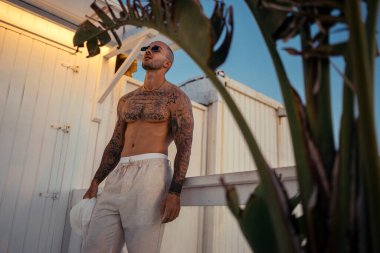 Beyaz gömlekli, dövmeli ve güneş gözlüklü yakışıklı genç adam çatıda dikiliyor ve başka tarafa bakıyor.