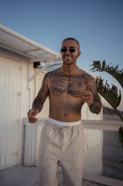 Sabahları sahilde poz veren dövmeli ve güneş gözlüklü yakışıklı genç bir adamın portresi.