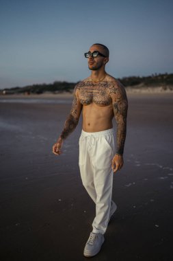Yazın kumsalda duran güneş gözlüklü ve dövmeli beyaz gömlekli yakışıklı genç adam.