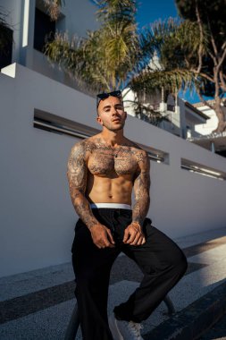 Kumsaldaki bankta poz veren dövmeleri olan yakışıklı, üstsüz genç bir adam..