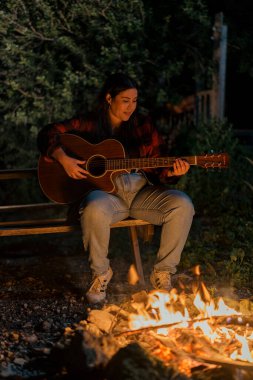 Genç bir kadın akşam kamp ateşinde gitar çalıyor.
