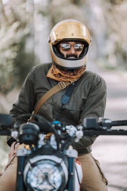 Kasklı ve gözlüklü genç sakallı adam motosikletin yanında oturuyor.
