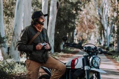 Şapkalı ve gözlüklü sakallı motorcu kırsalda sırt çantasıyla oturuyor.