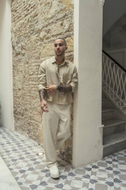 Beyaz takım elbiseli, şapkalı, yüzünde bir gülümseme ve bir otel odasının kapısında duran genç adam.