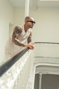 Beyaz gömlekli ve güneş gözlüklü yakışıklı genç adam merdivenlerde oturuyor ve kameraya bakıyor..