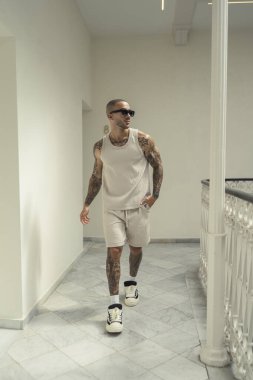 Dövmeli genç adam ayakta ve beyaz iç çamaşırlarıyla poz veriyor.