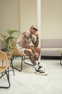 Beyaz elbiseli dövmeli bir adam bir sandalyede oturuyor ve dairede penceresi olan oturma odasında çay içiyor.