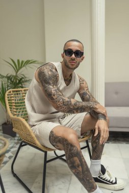 Yakışıklı, dövmeli, şık güneş gözlüklü genç adam odada kameraya poz veriyor.