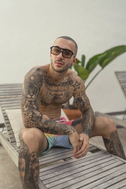 Dövmeli adam sahilde oturuyor.