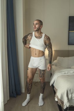 Genç, üstsüz, beyaz iç çamaşırlı, sakalsız adam yatak odasında duruyor.